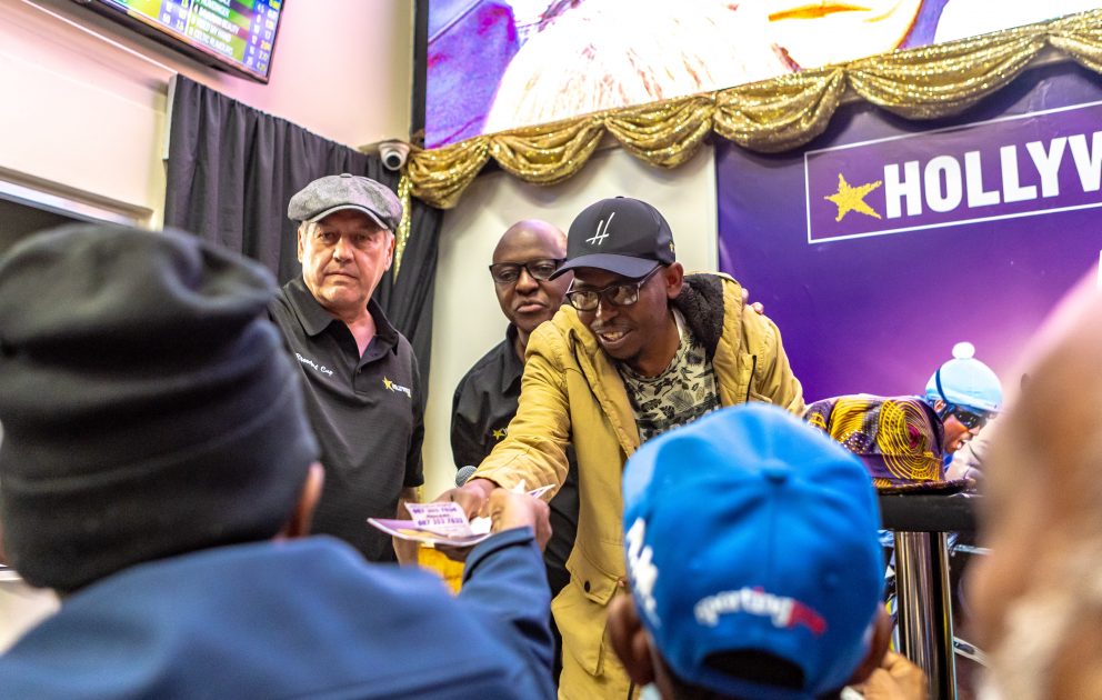 Sanele Mchunu hands a Hollywoodbets voucher to a happy punter at the Hollywoodbets Durban July pre-party at Springfield Park , as Owen Heffer and Brian Makwabarara look on 